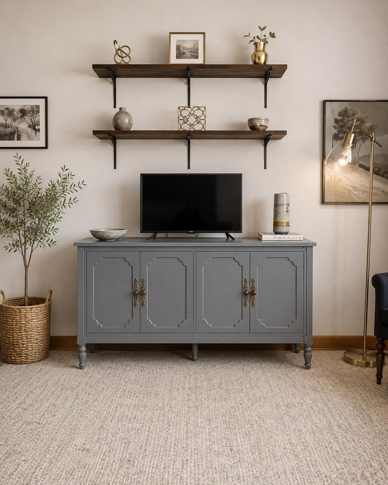 May include: A gray cabinet with four doors and ornate brass handles. A flat-screen TV sits on top of the cabinet. Two dark wooden shelves with decorative items are mounted on the wall above the cabinet.