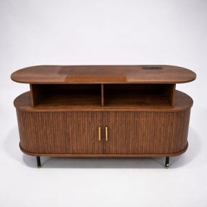 May include: A mid-century modern style wooden cabinet with a dark brown finish. The cabinet has a rounded shape with vertical grooved detailing on the front doors. It features two open shelves and gold-colored handles. The cabinet sits on black legs with gold accents.