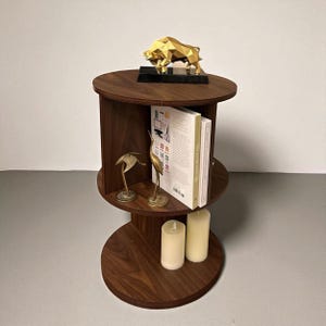 May include: A dark brown, three-tiered side table with a gold geometric bull statue on top. The middle shelf holds books and two brass bird figurines. Two cream-colored candles sit on the bottom shelf. The table is a stylish home decor item.