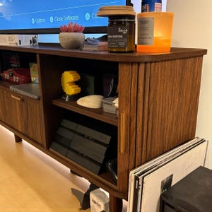 May include: A mid-century modern style wooden entertainment center with a fluted door design. The cabinet has open shelving and a closed cabinet with a gold handle. Items on display include a red toy car, a yellow Pac-Man figure, and a candle.