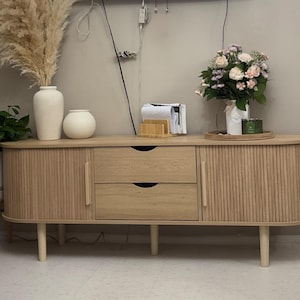 May include: A light wood-toned sideboard with two drawers and two doors. The doors have vertical ribbed detailing. The sideboard is on short, light-colored legs. Decorative items on top include vases, flowers, and a tray.
