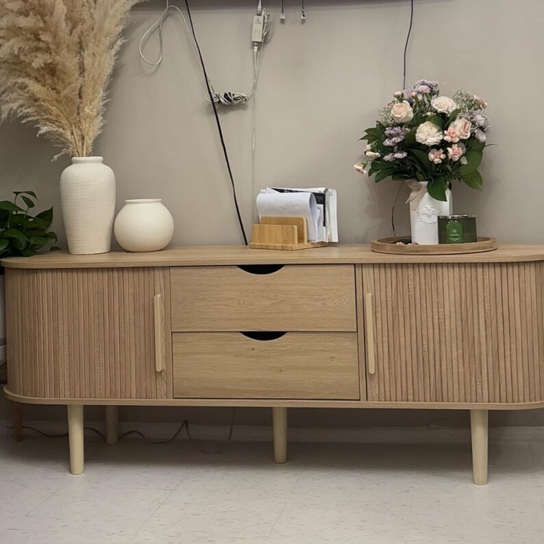 May include: A light wood-toned sideboard with two drawers and two doors. The doors have vertical ribbed detailing. The sideboard is on short, light-colored legs. Decorative items on top include vases, flowers, and a tray.