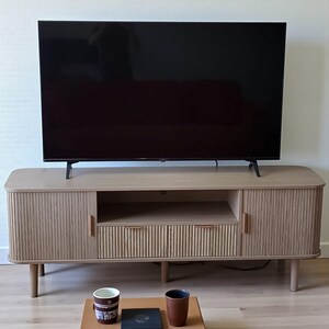 May include: A light-colored wooden TV stand with a large black television on top. The stand has a ribbed design on the doors and drawers, with a small open shelf. Two mugs and a small tray are on the floor in front of the stand.