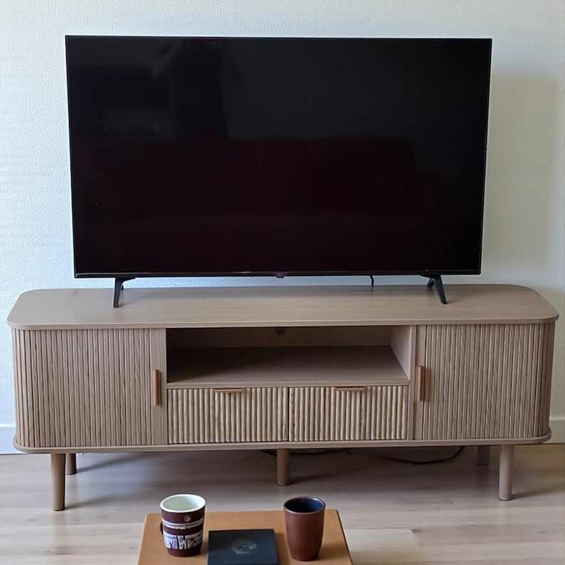 May include: A light-colored wooden TV stand with a large black television on top. The stand has a ribbed design on the doors and drawers, with a small open shelf. Two mugs and a small tray are on the floor in front of the stand.