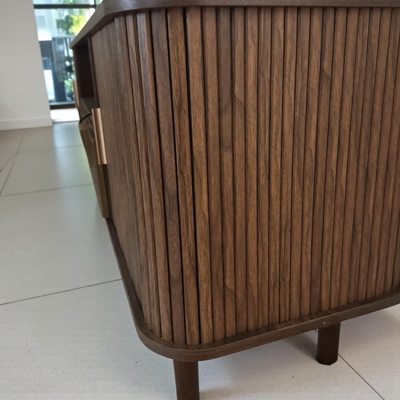 May include: A dark brown wooden cabinet with vertical grooved detailing. The cabinet has a curved base and legs, with a copper-colored handle. The wood grain is visible, and the overall design is modern and minimalist.