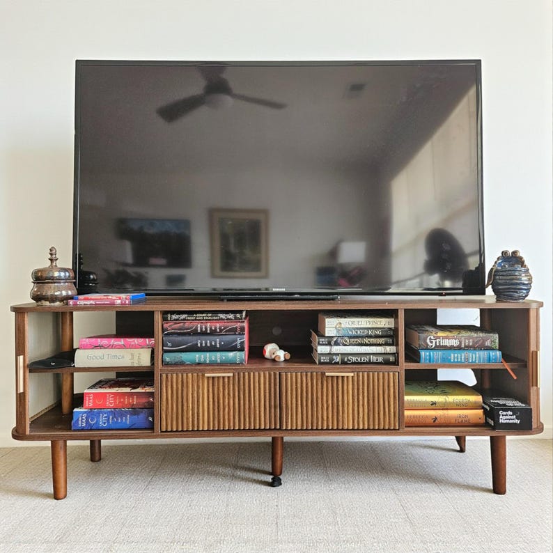 May include: A mid-century modern style TV stand in dark brown wood. The stand has a large open shelf, two drawers with vertical ribbed detailing, and angled legs. Books and decorative items are displayed on the shelves.