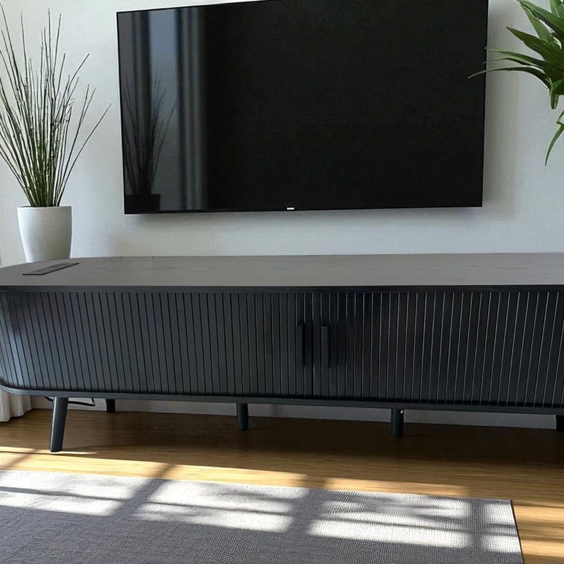 May include: A black entertainment center with a slatted front design, featuring a single door and a dark handle. The cabinet is set against a white wall with a large black television mounted above it. A gray rug sits on the wooden floor.