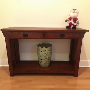 May include: A reddish-brown wooden console table with two drawers and a lower shelf. A green vase with a floral design sits on the shelf. A Santa Claus figurine stands on the table's surface.