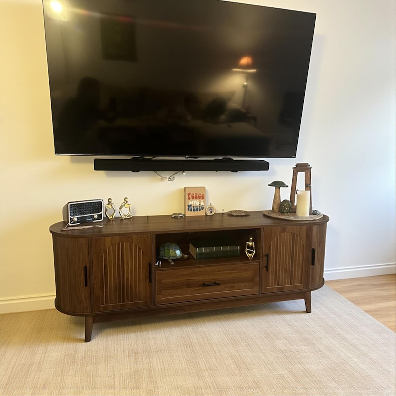 May include: A dark wood TV stand with a curved front and ridged door panels. The stand has a drawer and open shelf, with decorative items on top, including a radio, figurines, and a sign that reads "Raise Music". A large TV is mounted above.