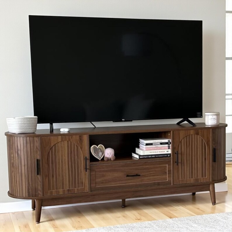 May include: A dark brown wooden TV stand with two doors, a drawer, and a center shelf. The doors have vertical ribbed detailing. A large black TV sits on top. Decorative items include books, a candle, and a white basket.