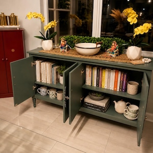 May include: A dark green cabinet with open doors, displaying books, mugs, and a teapot. The top of the cabinet features a decorative bowl, figurines, and yellow flowers in white geometric vases. The cabinet is set against a window.