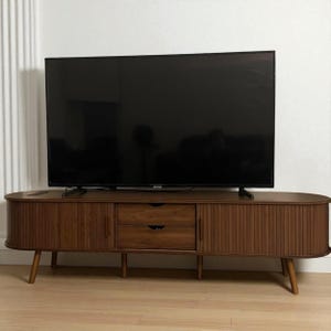 May include: A mid-century modern style TV stand in a warm brown finish. The stand features two drawers, two doors, and a slatted design on the doors. The stand is supported by four angled wooden legs and holds a large black television.