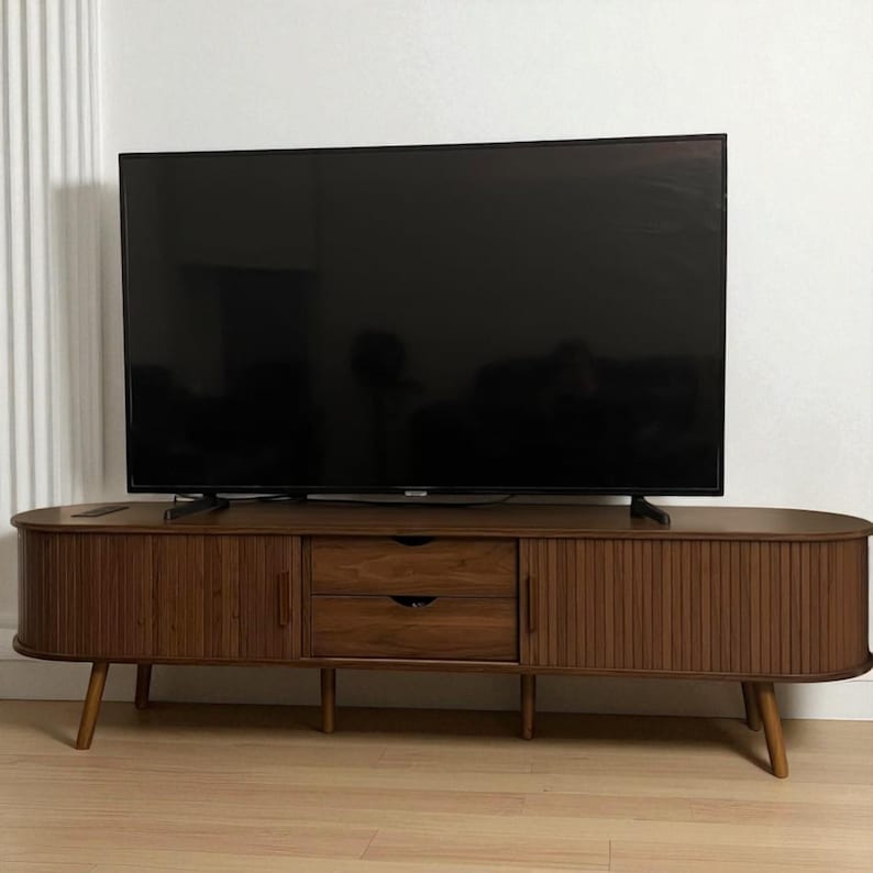 May include: A mid-century modern style TV stand in a warm brown finish. The stand features two drawers, two doors, and a slatted design on the doors. The stand is supported by four angled wooden legs and holds a large black television.