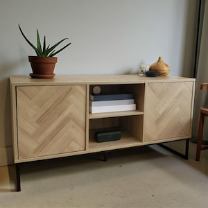May include: A light oak wood media console with a herringbone pattern on the doors. The console has black metal legs and an open shelf with books and a speaker. A potted plant and decorative items sit on top.