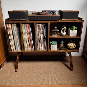 May include: A wooden record player console with a turntable, speakers, and storage for vinyl records. The console has a mid-century modern design with angled legs and multiple shelves. Decorative items like small plants and a perfume bottle are displayed.