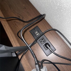 以下が含まれることがあります： 2つのUSBポートと2つの標準的な電気コンセントを備えた黒い電源コンセント。コンセントは木製の表面に埋め込まれており、いくつかの黒いコードが差し込まれています。木製の表面には茶色の木目模様があります。