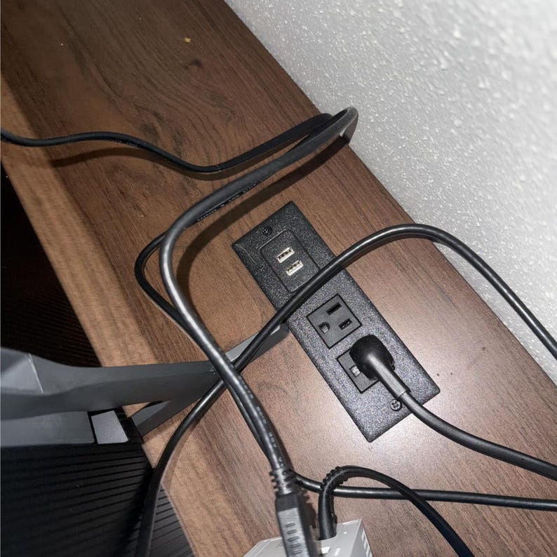 以下が含まれることがあります： 2つのUSBポートと2つの標準的な電気コンセントを備えた黒い電源コンセント。コンセントは木製の表面に埋め込まれており、いくつかの黒いコードが差し込まれています。木製の表面には茶色の木目模様があります。