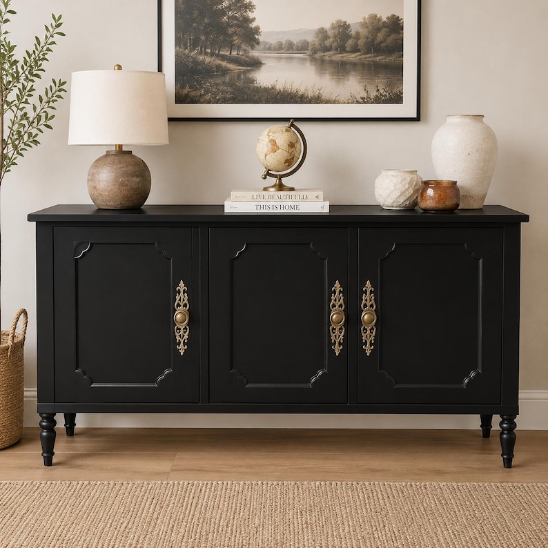 May include: A black sideboard with three doors and ornate gold hardware. The cabinet has a rectangular shape and sits on four short, turned legs. A lamp, globe, books, and vases are displayed on top. A framed landscape painting hangs on the wall.