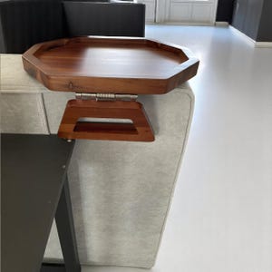 May include: A wooden sofa arm tray with a dark brown finish. The tray has an octagonal shape and is attached to a wooden armrest with a metal hinge. The armrest has a rectangular opening.