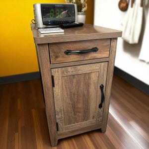 May include: A wooden nightstand with a drawer and a cabinet door. The nightstand has a light brown finish with black hardware. An alarm clock, remote, and small plant sit on top. The clock displays the time 8:32.