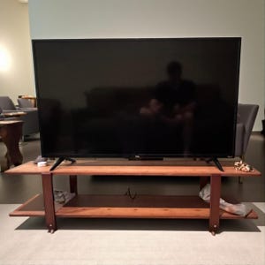 May include: A large, black flat-screen television sits atop a two-tiered, brown wooden media console. The console has a lower shelf and is supported by dark brown legs. The TV is centered on the console, with a reflection on the screen.
