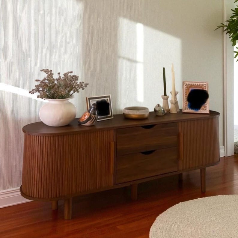 May include: A mid-century modern style wooden cabinet with two drawers and two doors. The cabinet is a warm brown color and has a curved front with vertical grooves. Decorative items on top include a vase with dried flowers, candles, and framed art.