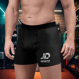 May include: Black athletic compression shorts featuring a wide elastic waistband and the white text JD SPORTS UNLEASH YOUR POWER printed on the lower left leg. The shorts are constructed from a smooth, form-fitting fabric with visible seams.