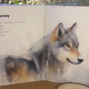 May include: An open book featuring a watercolour illustration of a grey wolf with brown accents and snow on its fur. The text reads: Journey. The wolf's howl echoes a timeless truth: Run wild. Live deep. Fear may follow. Keep going.