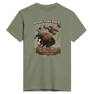 May include: An olive green t-shirt featuring a graphic of a bison tossing a person into the air. The text reads BISON TOSS DAYS, The Humor Is Fiction. The Danger Is Real. YELLOWSTONE. The illustration shows a person in a red plaid shirt and blue jeans.