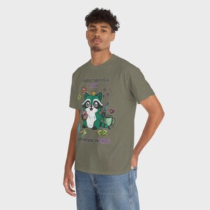 May include: Olive green t-shirt featuring a cartoon raccoon wearing a crown and holding a donut. The text on the shirt reads "PRODUCTIVITY IS A MYTH, I'M THRIVING IN CHAOS."