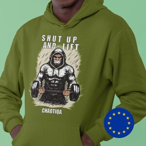 Può includere: Felpa con cappuccio verde oliva con la scritta "SHUT UP AND LIFT" sopra una grafica di un sollevatore di pesi. La parola "CHAOTIQA" è sotto la grafica. La felpa ha un cappuccio con coulisse e una tasca frontale.
