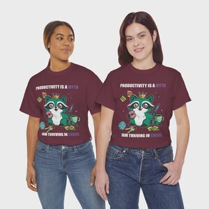 May include: Two maroon t-shirts with the text "PRODUCTIVITY IS A MYTH" and "IAM THRIVING IN CHAOS". Each shirt features a cartoon raccoon wearing a crown and holding a donut. The raccoon is surrounded by office supplies.