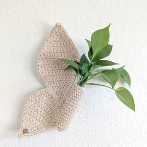 May include: A beige crochet wall planter shaped like a butterfly wing, with a small wooden button detail. The planter holds a green leafy plant. The wall decor is mounted on a textured white wall.