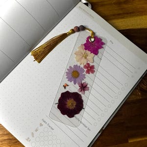 Può includere: Un segnalibro rettangolare trasparente con fiori pressati incorporati nelle tonalità magenta, rosa chiaro e viola. Il segnalibro include un occhiello in metallo dorato attaccato a una nappa in seta dorata e due piccole perline scure in cima.