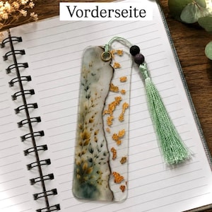 Könnte beinhalten: Ein rechteckiges Lesezeichen aus Kunstharz mit geteiltem Design, das auf einer Seite einen botanischen Druck mit grünen und gelben Blättern und auf der anderen Seite Blattgoldflocken zeigt. Es hat eine mintgrüne Quaste und zwei dunkle Perlen.