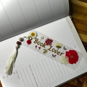 Può includere: Un segnalibro rettangolare trasparente con fiori pressati rossi, bianchi e gialli e piccoli dettagli a forma di farfalla. La scritta "Book Lover" è stampata in oro. È presente una nappa bianca con perline grigie e rosse fissata in alto.