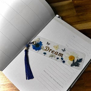 Könnte beinhalten: Ein transparentes, rechteckiges Lesezeichen mit gepressten blauen und weißen Blumen, kleinen Schmetterlingsmotiven und dem Wort Dream in goldener Schrift. Es hat eine goldene Metallöse mit einer dunkelblauen Quaste und einer kleinen Perle.