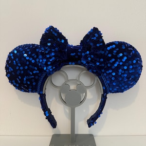 May include: A headband featuring two round ears covered in dark blue sequins and a central dark blue sequined bow. The headband is displayed on a grey stand with the text EarsBYNatalie printed on the base, showcasing a consistent blue design.