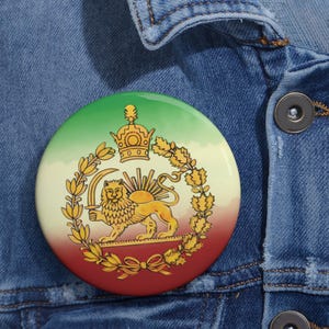Op de afbeelding: Een ronde button met een gradiënt achtergrond van groen, wit en rood. De button toont een gouden leeuw met een zwaard, een kroon en een zonnesymbool, omringd door een gouden krans. De button is geplaatst op een spijkerjasje.