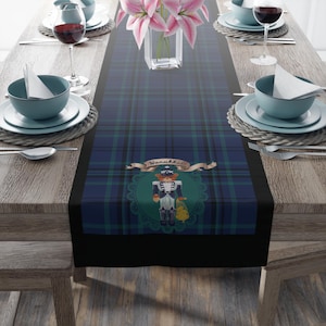 May include: A table runner with a blue and green plaid pattern, featuring a decorative nutcracker design with the word "Hanukkah" on a banner. The table is set with plates, bowls, silverware, and wine glasses.