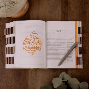May include: An open book with the words "And You Will Be My Witnesses" in orange script. The word "acts" is visible on the right page. A gray pen rests on the pages. Book tabs and a globe are also visible.