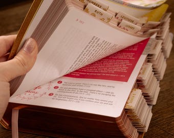May include: A gold-edged Bible with labeled tabs for easy navigation. The open pages reveal text and a pink ribbon bookmark. The tabs are labeled with book names like Numbers, Ruth, and Leviticus. The Bible is open on a wooden surface.