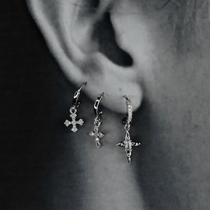 Puede incluir: Tres pendientes de plata con colgantes de cruz. Los pendientes tienen diferentes diseños, uno con forma de cruz adornado con pequeñas piedras brillantes. Los otros dos pendientes también tienen colgantes de cruz, uno con un diseño más detallado.