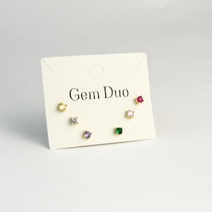 Puede incluir: Un conjunto de seis pendientes de botón con gemas de colores, presentados en una tarjeta crema. Los pendientes tienen engastes dorados e incluyen gemas amarillas, transparentes, moradas, verdes, rosas y rojas. La tarjeta lleva el texto "Gem Duo".