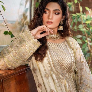 May include: A woman wearing a white and gold embroidered dress with a delicate lace trim. The dress has a sheer overlay and intricate patterns. She is wearing pearl jewelry and gold earrings.