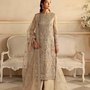 Puede incluir: Una mujer lleva un vestido largo de color crema con intrincados bordados plateados y un dupatta transparente a juego. El vestido tiene un escote redondo y mangas largas. El dupatta está drapeado sobre sus hombros y está decorado con un borde plateado.