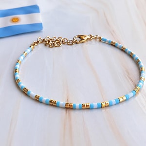 May include: A delicate bracelet featuring alternating light blue, white, and metallic gold cylindrical beads arranged in a repeating pattern. The bracelet includes a gold-toned metal lobster clasp and an adjustable chain link extension.