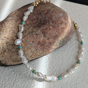 Può includere: Un delicato braccialetto con perle bianche irregolari, piccole perline turchesi e dettagli dorati. Il braccialetto è esposto su una superficie di pietra strutturata, che ne mette in risalto l'elegante design. La chiusura è dorata.