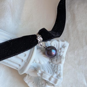 Könnte beinhalten: Ein schwarzes Samt-Choker-Halsband mit einem dunkel irisierenden Perlenanhänger. Der Anhänger ist mit einem silberfarbenen, blattförmigen Verschluss am Choker befestigt. Die Halskette wird auf einem weißen Dekorationsgegenstand präsentiert.