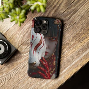 May include: A smartphone case featuring a digital art design. The case displays a woman holding a white fox mask with red accents. The woman has dark hair and is wearing a red and gold outfit. The case has a black background.